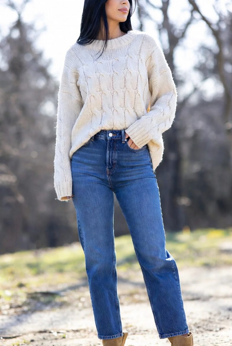 Stylish slim woman wearing high-waist straight blue jeans with a cream cable-knit sweater and tan boots, walking confidently on a winter street.
