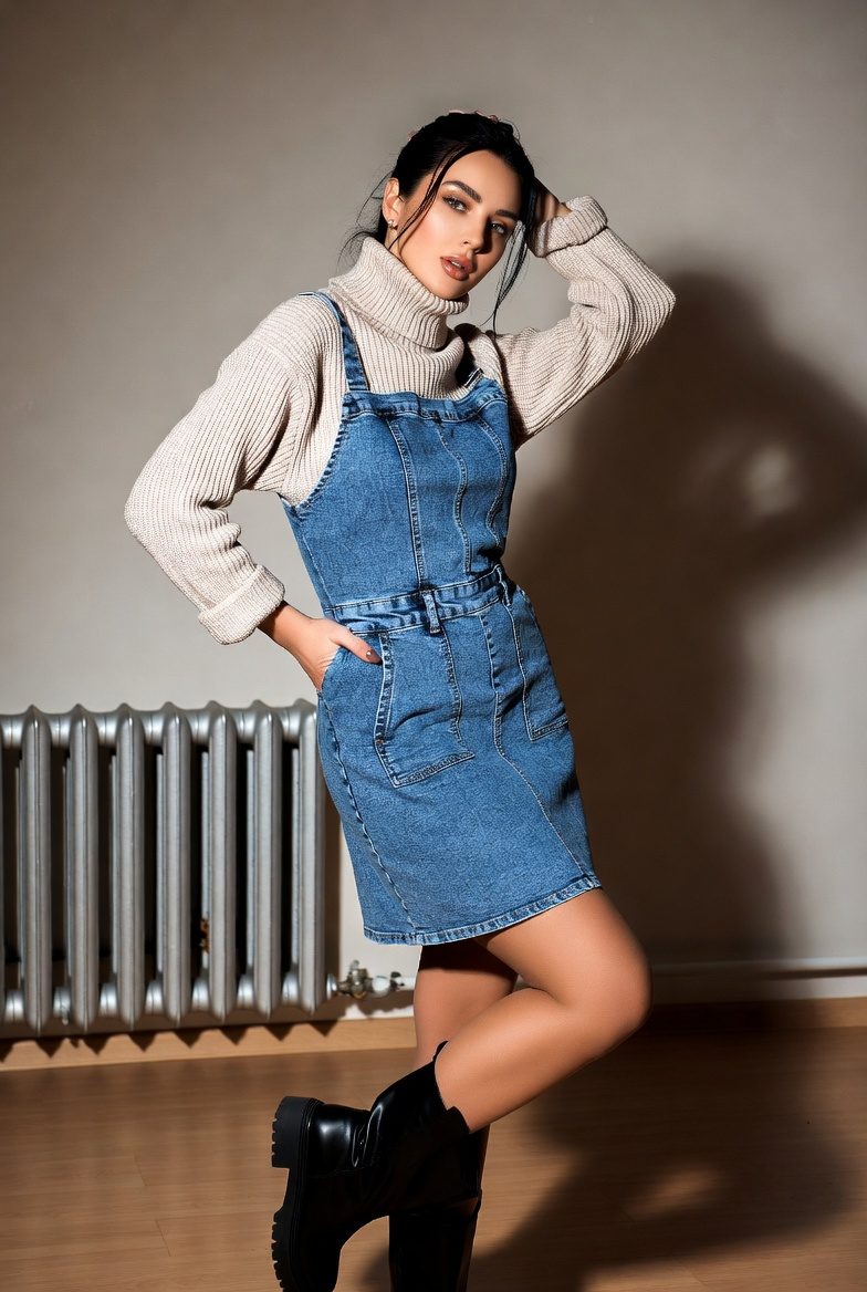 Petite slim woman wearing a sleeveless denim dress layered over a beige turtleneck and tall boots indoors.