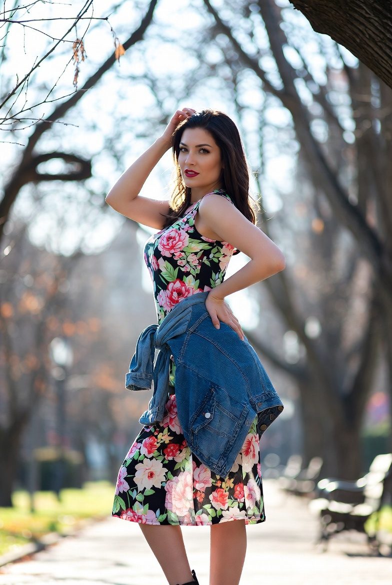 Curvy woman in floral midi dress and denim jacket posing in a December park with ankle boots.