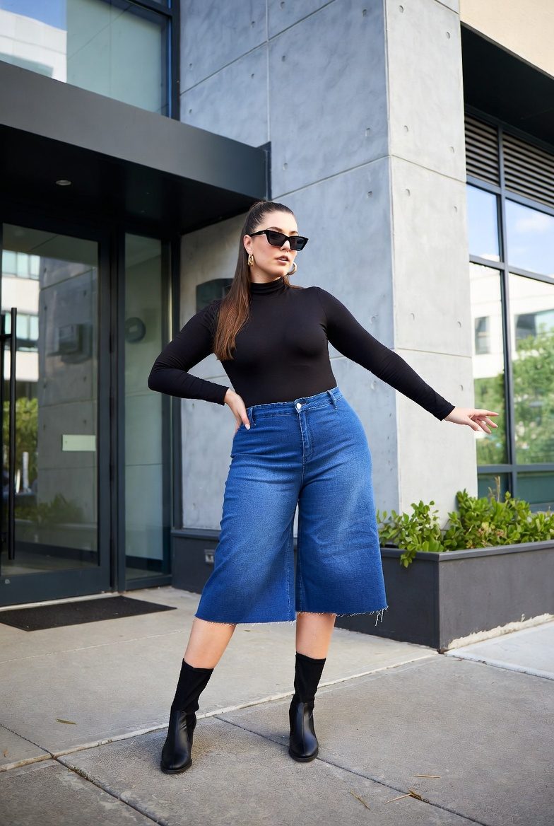 Plus-size woman in cropped denim culottes, black turtleneck, and knee-high boots posing in an urban backdrop.
