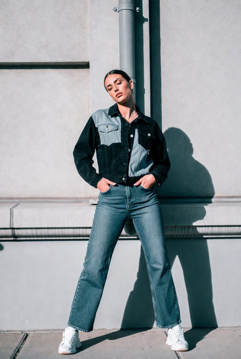 Slim woman wearing a patchwork denim jacket with black jeans and sneakers posing casually by an urban wall.