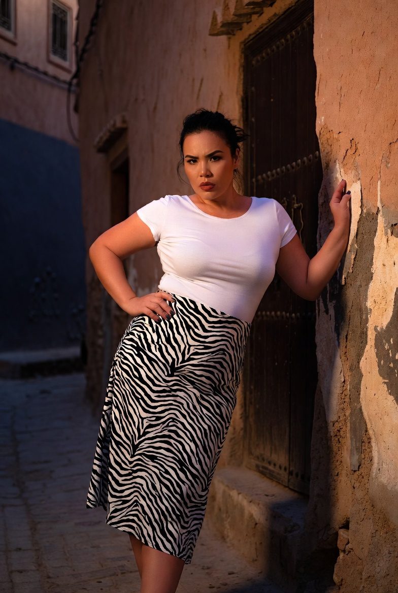 Medium-built Asian woman in zebra-print midi skirt and white tee leaning against Moroccan street wall, casual-chic travel outfit.