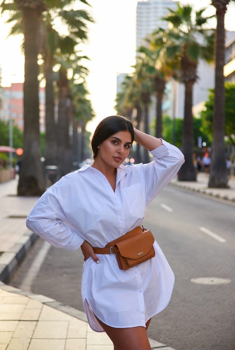 Medium-built brunette woman in white oversized shirt dress cinched with tan belt bag on city street with palm trees, casual vacation look.