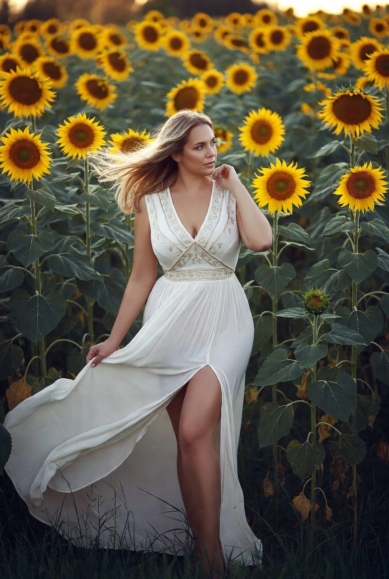 Curvy white woman wearing flowing embroidered boho maxi dress in sunflower field, golden light, romantic vacation style.