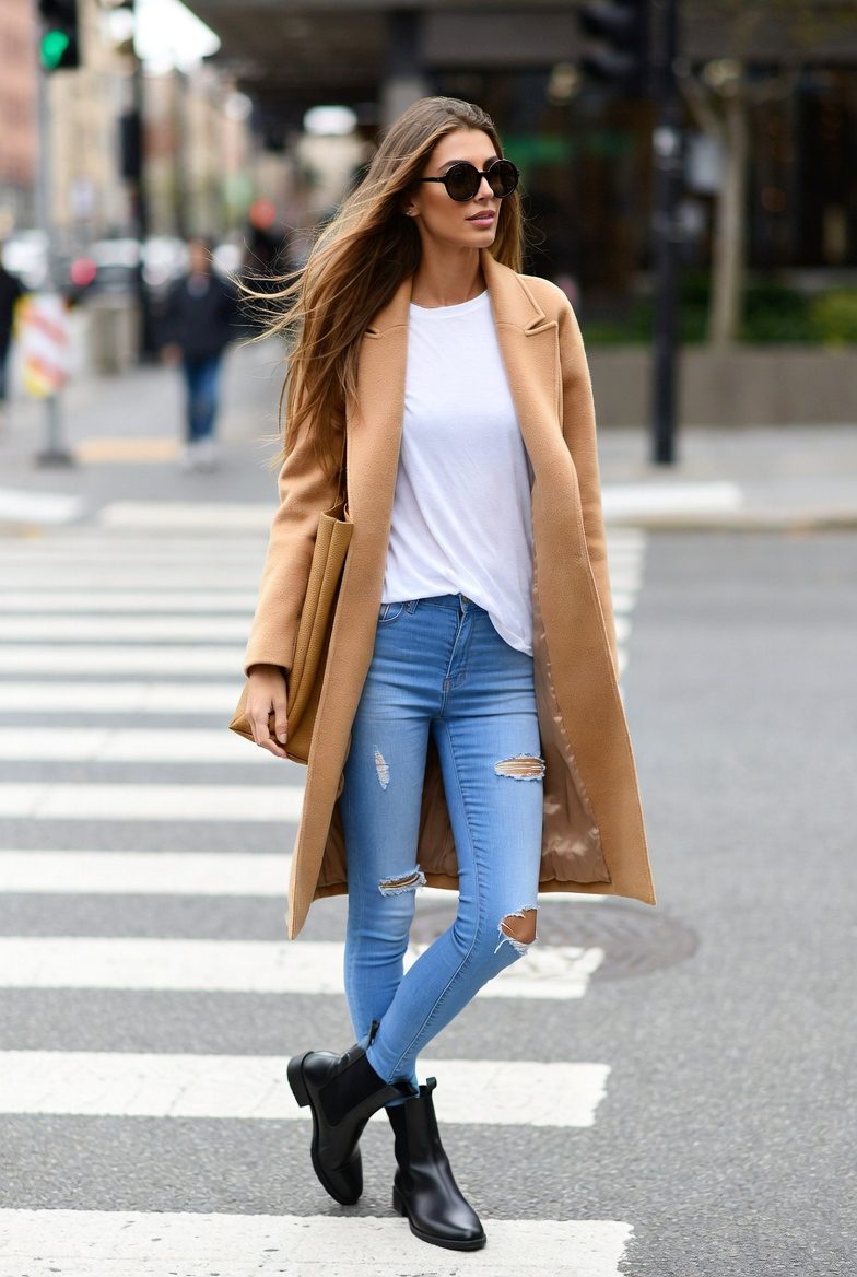 Slim white woman in a camel coat, white T-shirt, ripped skinny jeans, and Chelsea boots walking through a fall city street.
