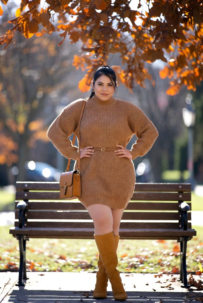 Latina woman wearing a camel knit sweater dress with belt and tan knee-high boots sitting on a park bench surrounded by fall leaves.