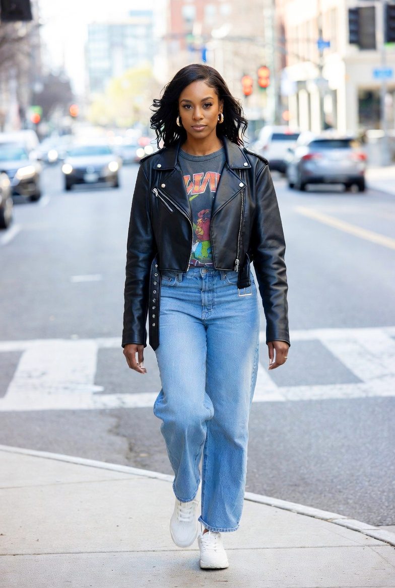 Black woman wearing a cropped leather jacket, graphic T-shirt, boyfriend jeans, and white sneakers in a downtown autumn setting.