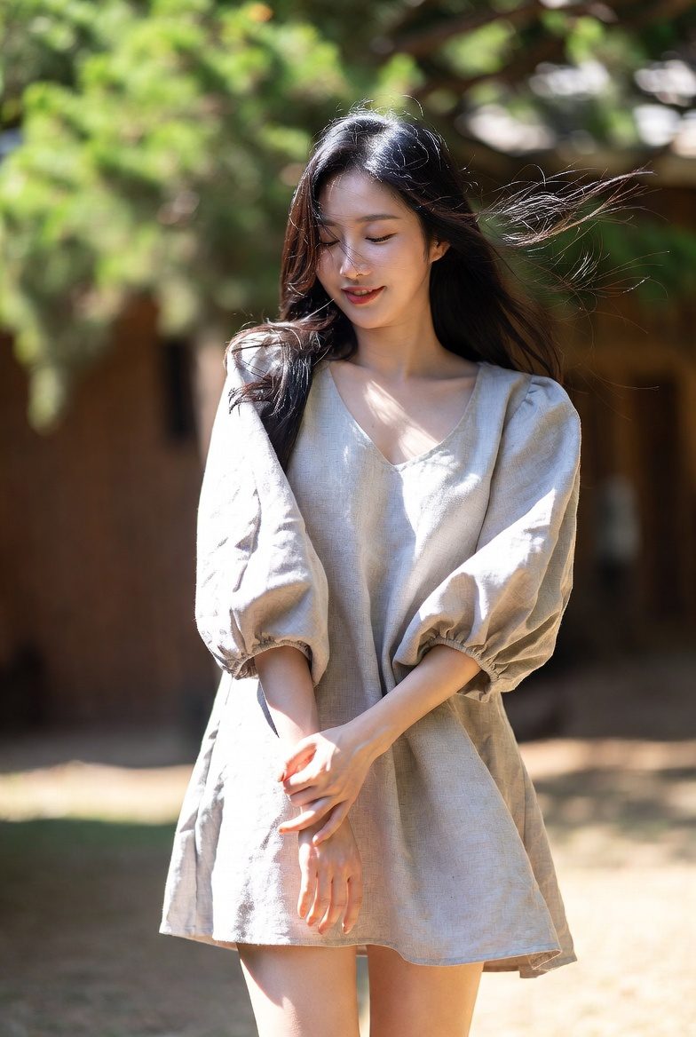 Korean woman wearing a linen mini dress with balloon sleeves, standing in warm natural sunlight.