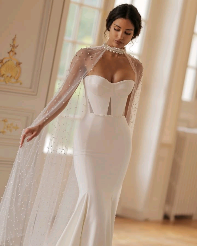 Elegant white wedding dress with a fitted bodice, pearl-embellished sheer cape, and a flowing mermaid skirt, worn by a woman with an updo hairstyle, standing in a luxurious room with large windows and ornate decor.